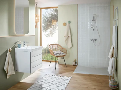 Łazienka z umywalką, dużym lustrem i prysznicem z kolekcji O.novo Nowoczesna łazienka z umywalką ścienną, dużym lustrem, wiklinowym krzesłem i białym prysznicem. Na haczykach wiszą ręczniki w neutralnych kolorach, a pomieszczenie oświetla naturalne światło wpadające przez duże okno.