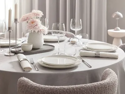 Runder Esstisch mit weißen Afina Tellern, Afina Gläsern, Besteck, Servietten und einer Vase mit rosa Blumen.