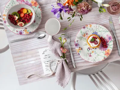 Une table décorée de motifs floraux avec des assiettes, des couverts, une serviette, un petit dessert et une coupe de fruits sur une nappe claire.