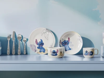 A set of plates and cups from the Disney Stitch collection featuring cartoon motifs are displayed on a light blue shelf with utensils.