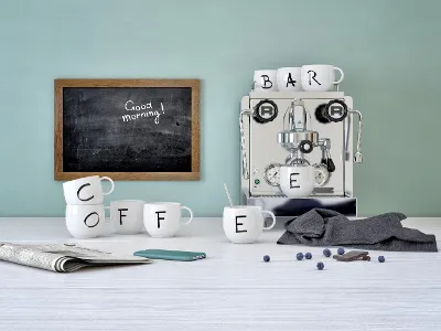 Espressomaschine mit Villeroy & Boch „Letters“-Tassen, die das Wort „COFFEE“ buchstabieren, auf einem Tisch, neben einem Telefon, einer Zeitung, Blaubeeren und einer Tafel mit der Aufschrift „Guten Morgen!“.