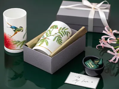 Präsentiert werden zwei Amazonia Tassen mit Blumen- und Vogelmotiven Präsentiert werden zwei Amazonia Tassen mit Blumen- und Vogelmotiven, eine Geschenkbox, eine kleine Schale mit eingepackten Artikeln und eine rosa Schleife auf grünem Untergrund.