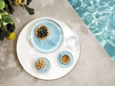 A round tray with tableware from the Perlemor Aqua collection and a glass of water, at the edge of a pool.