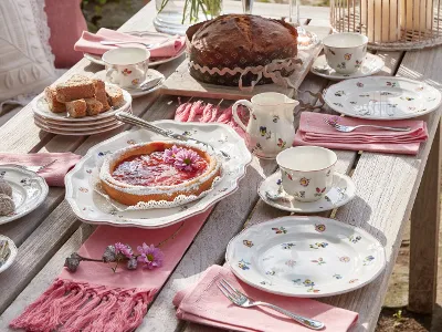 Die Petite Fleur-Kollektion von Villeroy & Boch ist elegant auf einem Holztisch arrangiert und umfasst Teller mit Blumenmuster, einen Kuchen, eine Torte, Tassen und rosa Servietten zum Tee.