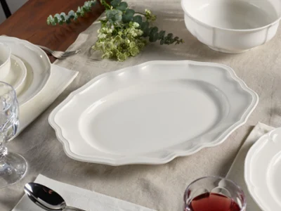 A white platter with scalloped edges is elegantly placed on a set dining table adorned with a tablecloth, surrounded by plates, glasses, cutlery, and bowls.