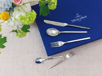 Besteckset mit Messer, Löffel und Gabel auf blauem Tuch neben Blumenarrangement auf beigem Untergrund.	