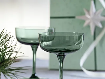 Deux verres à cocktail verts sur une surface blanche avec une boîte cadeau verte et des branches de sapin en arrière-plan.