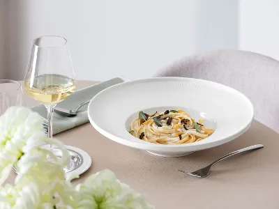 An Afina plate with pasta, mushrooms and herbs, accompanied by a glass of white wine on an elegantly laid table.