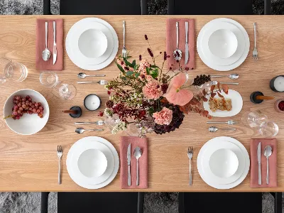 A wooden dining table with white plates, bowls, cutlery on pink napkins, two candles, a bowl of grapes, and a floral arrangement.