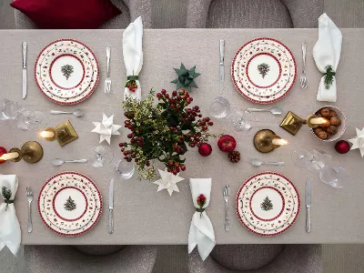 Une table de salle à manger dressée pour quatre personnes avec une décoration festive, dont des assiettes Toys Delight avec des motifs de sapin de Noël, des notes rouges et dorées, des serviettes blanches pliées, des bougies et un centre de table avec des baies rouges et des plantes vertes.