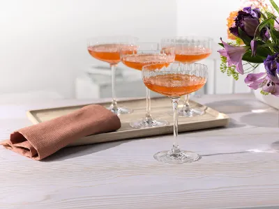Rose Garden champagne coupe glasses Four glasses filled with orange cocktails on a tray stand next to a folded napkin and flowers on a light-colored table.