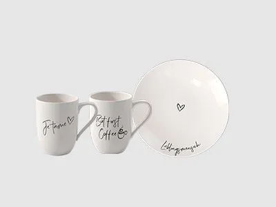 Zwei weiße Tassen und ein weißer Teller mit schwarzem handschriftlichem Text und einer kleinen Herzillustration auf jedem Artikel.