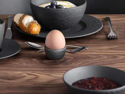 A breakfast set with coffee, juice, a croissant, jam and a soft-boiled egg on a wooden table, green flowers are visible.