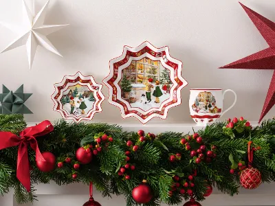 Une présentation aux couleurs de Noël se composant de deux assiettes « Toy’s Fantasy » de Villeroy & Boch, d’une tasse « Toy’s Fantasy », d’étoiles en papier et d’une abondante guirlande de rubans rouges, d’ornements et de baies sur un manteau de cheminée blanc.