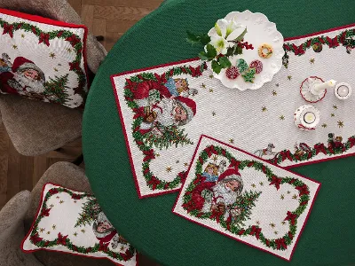 Toy's Fantasy Accessoires auf einem Tisch mit grüner Tischdecke Ein runder Tisch mit einer grünen Tischdecke ist mit weihnachtlichen Tischsets und Kissen aus der Toy's Fantasy Kollektion mit Weihnachtsmannmotiven dekoriert. Auf dem Tisch stehen außerdem Kerzen und ein dekorativer weißer Teller.
