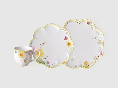 Auf einem schlichten Hintergrund sind eine Tasse mit Blumenmuster und zwei passende Teller mit Wellenrand und grünen und gelben Akzenten zu sehen.