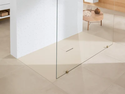 Prysznic bezprogowy Squaro Infinity w łazience z beżowymi płytkami podłogowymi Minimalistyczna łazienka z beżowymi płytkami podłogowymi, prysznicem zlicowanym z podłogą oddzielonym szklanym panelem, ścianą wyłożoną białymi kafelkami i małym drewnianym stolikiem z elementami dekoracyjnymi w tle.