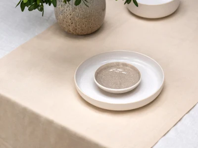A table with a beige runner, a stack of ceramic plates, a bowl, and a vase with green leafy branches.

