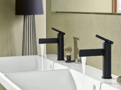 Ein modernes Badezimmer mit  zwei schwarzen Wasserhähnen über einem weißen Waschbecken. Auf der Ablage liegen ein Rasierpinsel und ein Handtuch. 