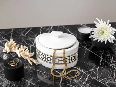 Eine dekorative, gold-gemusterte MectroChic Zukderdose auf einer Marmoroberfläche, umgeben von getrockneten Korallen, einer Kerze, einer Blume und einem Ring auf einem schwarzen Deckel.