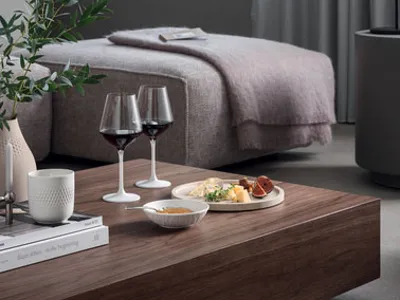 Modern bedroom with a cozy bed, large window, and gray curtains. A wooden table holds books, Manufacture wine glasses, a plate of cheese and snacks, and a potted plant in the foreground.