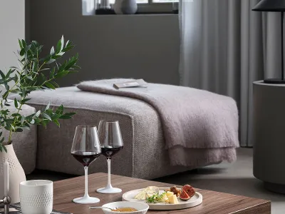 Modernes Schlafzimmer mit gemütlichem Bett, großem Fenster und grauen Vorhängen. Im Vordergrund ein Holztisch mit Büchern, Manufacture Weingläsern, einer Käseplatte, Snacks und einer Topfpflanze