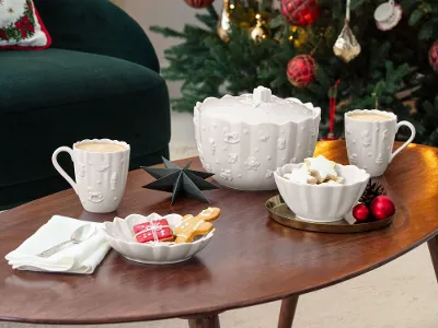 Un sapin de Noël décoré d'ornements rouges se dresse derrière une table basse ornée de tasses, de biscuits et de décorations festives dans un salon confortable.