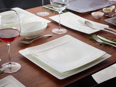 The Modern Grace dining set from Villeroy & Boch consists of square plates, wine glasses, bowls, and flatware arranged on a wooden surface with fern accents. A glass of red wine complements the ambience perfectly.