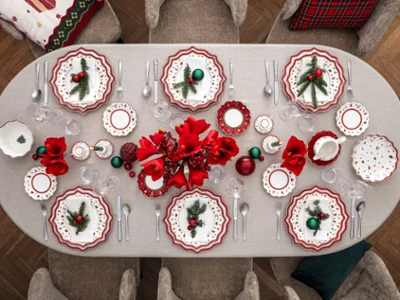 Una mesa de comedor para ocho comensales decorada festivamente con platos rojos y blancos Toy's Delight de Villeroy & Boch, cubertería en orden, flores rojas, adornos navideños y un mantel gris en un acogedor comedor con asientos grises.