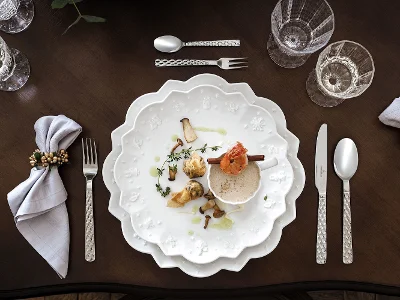 Une table de salle à manger sophistiquée présente les assiettes « Boston » de Villeroy & Boch, artistiquement décorées de motifs ondulés, et complétées par des verres en cristallin, de couverts de table argentés richement décorés et de serviettes décoratives sur une table en bois foncé.