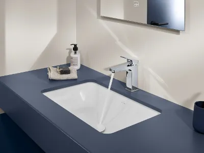 Ein modernes Waschbecken mit fließendem Wasser, einem Seifenspender, einem Waschlappen mit einem Stück Seife und einem Spiegel darüber. Die Ablage ist blau und rechts neben dem Waschbecken befindet sich ein Zahnbürstenhalter.