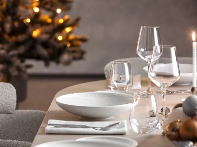  Une table à manger dressée avec des verres et des assiettes Afina, à côté une bougie allumée et un sapin de Noël décoré en arrière-plan.
