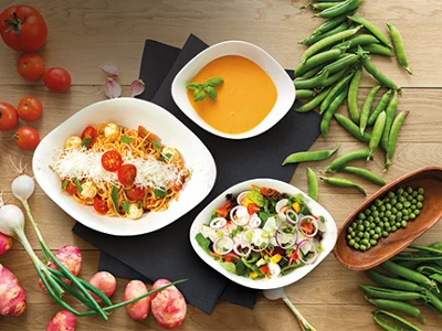 Set básico Vapiano de Villeroy & Boch sobre una mesa de madera con tomates, cebolletas, guisantes, judías y cebollas. Plato Vapiano relleno de pasta, ensalada y sopa de calabaza 