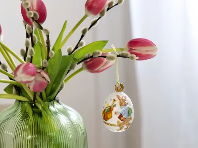 Un bouquet de tulipes et de chatons d'osier dans un vase vert, orné d'un délicat œuf décoratif de l'édition annuelle de Pâques de Villeroy & Boch, suspendu à une branche. La lumière du soleil pénètre par la fenêtre à l'arrière-plan.