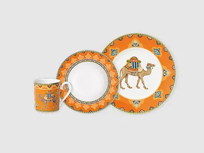 Set aus orangefarbenem Essgeschirr mit Kamelmuster, bestehend aus einer Tasse, einem Salatteller und einem Essteller auf grauem Hintergrund.