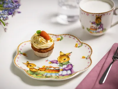 A carrot cupcake on an Annual Easter Edition 2024 plate with a bunny pattern, together with a glass of milk and a bouquet of grape hyacinths on a white tablecloth.