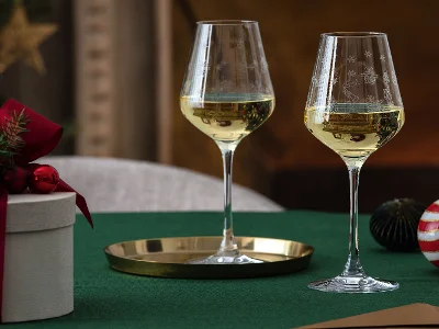 Deux verres à vin blanc sur une nappe verte sont accompagnés d’une boîte cadeau ronde blanche Toy's Delight de Villeroy & Boch avec un ruban rouge et une décoration festive.