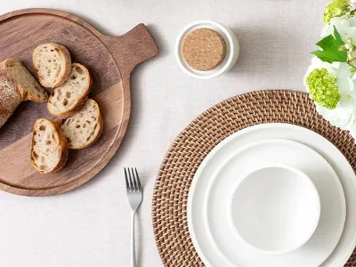 A table set with white dinnerware on rattan placemats with a wooden serving tray with bread on it