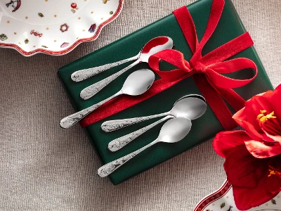 A green box with a red bow with five silver spoons, pictured on a gray background with a red flower and partially visible decorative plates from the Toy’s Delight collection by Villeroy & Boch.