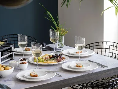 Ein Esstisch, gedeckt mit weißen NewMoon Tellern, Weingläsern, Käse, Obst und Vorspeisen, bereit für eine Mahlzeit.