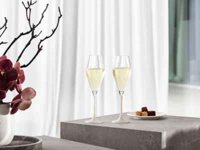 Dos copas de champán Manufacture con tallo blanco sobre una superficie gris, junto a un plato de bombones y un jarrón con flores rosas y ramas.