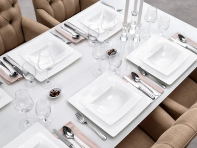 Ein hübsch gedeckter Esstisch mit weißem, quadratischem Geschirr der Marke Modern Grace von Villeroy & Boch, Silberbesteck, Weingläsern und beigen Servietten. Beige gepolsterte Stühle umgeben den Tisch. In der Mitte stehen drei Kerzenständer.