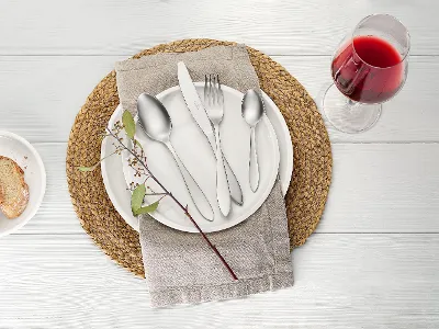 Ein gedeckter Tisch mit einem Artesano Teller, Arthur Besteck und einer Serviette auf einem runden Tischset. Daneben stehen ein Glas Rotwein und ein kleiner Teller mit Brotscheiben. Auf dem Teller liegt ein Zweig.