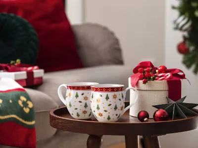 Zwei Weihnachtstassen, eine Geschenkbox und Weihnachtsschmuck auf einem kleinen Tisch neben einem festlich geschmückten Wohnzimmer.