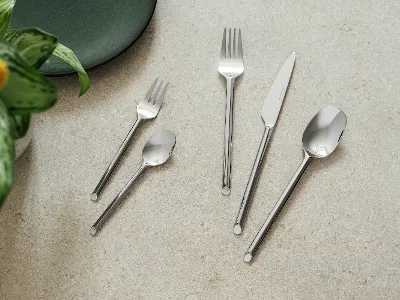 Fünf like. Besteckteile von like. by Villeroy & Boch – Gabel, Messer, Löffel, Dessertgabel und Teelöffel – sind auf einer hellen Steinoberfläche neben einem grünen Teller und einer Pflanze arrangiert.	