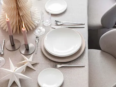 A beautiful dining table setting with white and beige NewMoon plates, cutlery, glasses, two lit candles and decorative paper stars on a light-coloured tablecloth.