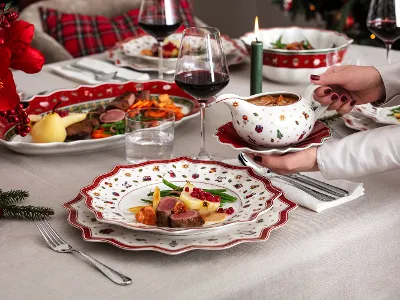 Ein festlicher Tisch ist mit weihnachtlichem Geschirr von Villeroy & Boch Toy's Delight gedeckt und präsentiert Teller mit Fleisch, Gemüse und Soße. Eine Person hält eine Sauciere, dazu Gläser mit Rotwein und eine brennende Kerze.