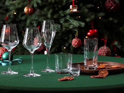 A stylish arrangement from the Toys Delight glassware collection by Villeroy & Boch, consisting of wine glasses and drinking glasses, elegantly placed on a green table. Dried citrus slices lie on a wooden tray in the foreground, complemented by the festive presence of a decorated Christmas tree in the background.
