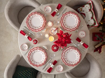Top-down view of a round dining table elegantly set for Christmas with Villeroy & Boch's Toy's Delight porcelain plates, red napkins, candles, and a red poinsettia centerpiece next to a lit Christmas tree.