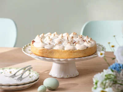 Easter Delight cake stand with Easter decorations A cake with meringue stands on a wooden table on a cake stand with Easter decorations from Easter Delight, next to plates and flowers.